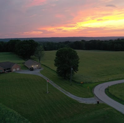 Aerial view of a country home surrounded by open fields, framed by a beautiful sunset in the sky