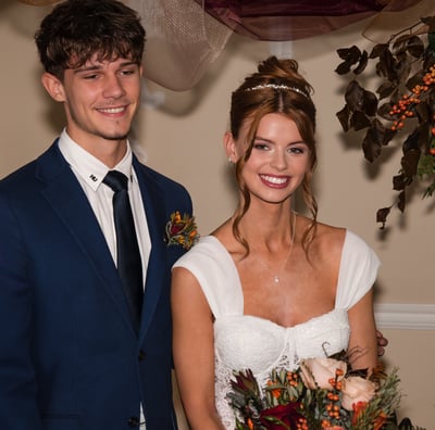 Smiling bride and groom pose indoors with floral bouquet and autumn-themed decor.