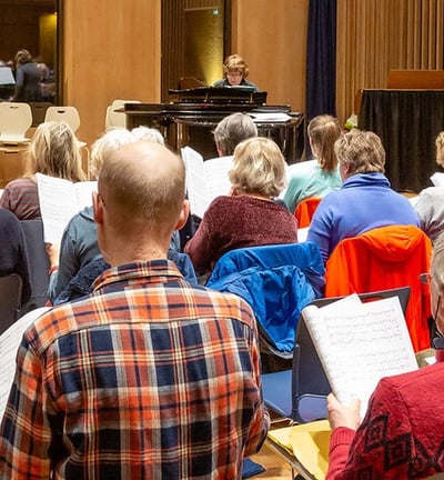 A choir is viewed from behind holding sheet music with a conductor in front of them behind a piano