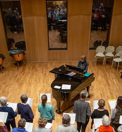 A choir can be seen from above holding sheet music. There is a grand piano in front of them.