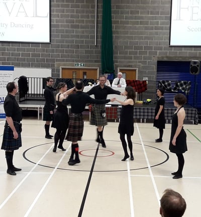 Dancers, dressed mostly in black, dancing a strathspey circle. Judges in the background on a stage.