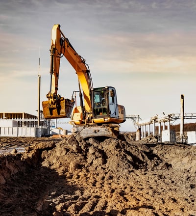 Bagger hebt Erde auf einer Baustelle aus bei Sonnenuntergang, im Hintergrund Rohbau und Kranstruktur