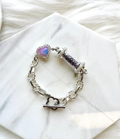 Features a silver chain with larger, oval links. It has a central cylindrical element with a dark pa
