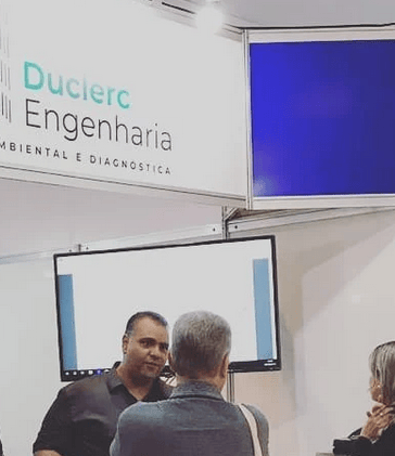 Representante da Duclerc Engenharia atendendo cliente durante feira técnica, apresentando serviços d