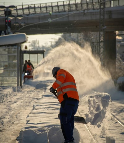 A crew of men in high visibility clothing snowblow and shovel snow