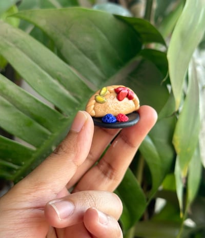 Clay gujiya-shaped fridge magnet held against a green leaf backdrop