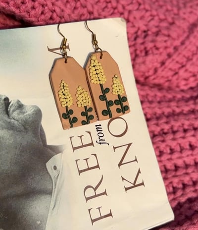 a pair of handmade yellow flower earrings displayed on a white book with pink cloth backdrop