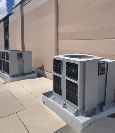 A series of air conditioning units are mounted on a metal structure outside a building with white paneling. Two large windows are visible behind the units, revealing the indoors with fluorescent lighting.