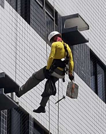 Técnico alpinista realizando teste de percussão e remoção de entulho em fachada predial, durante ins
