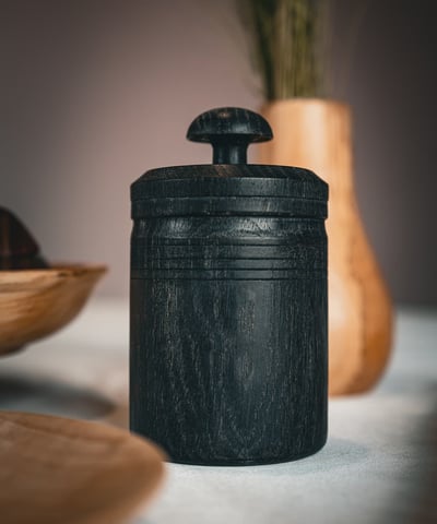 Pot à épices en bois foncé artisanal avec couvercle sculpté, posé sur une table. Sa texture naturell