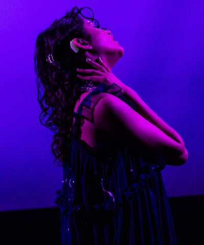 A brown woman with flowing hair and visible hearing aids, against a blue backdrop, bathed in pink