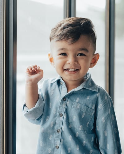 An adorable young boy standing in front of a window with his hand raised