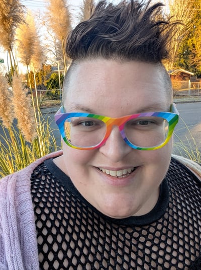 A knitter with short brown hair and rainbow glasses smiling.