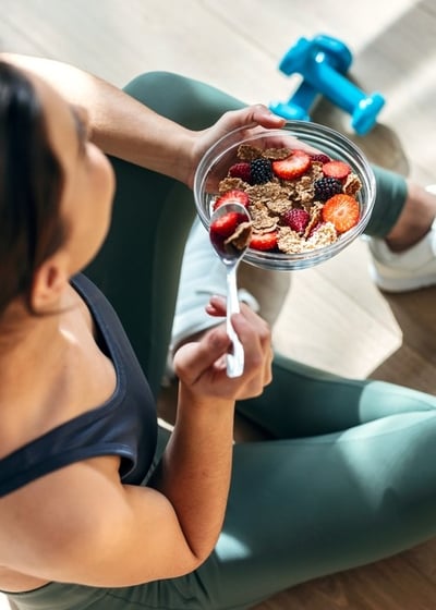 A woman in gym clothes sitting on the floor of a studio with a pair of weights, eating a healthy bowl of fruit and cereal