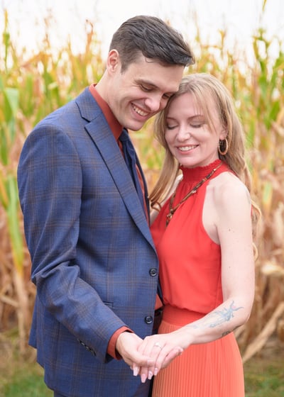 a man in a blue suit and woman in an orange dress holding hands in front of a corn field