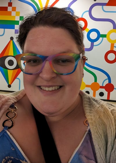 Image of a knitter smiling with rainbow glasses in front of a rainbow painting.