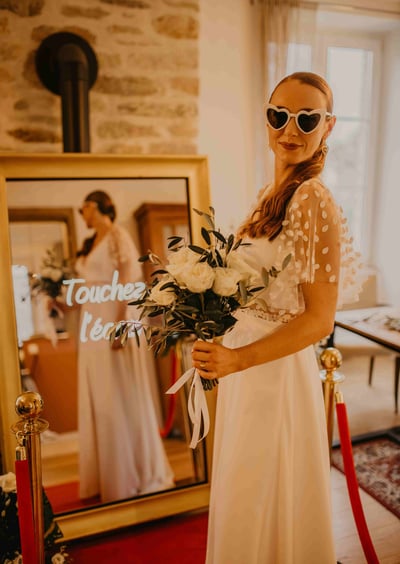 Une femme en robe de mariée qui pose devant le photobooth de pixtoevent avec des fleurs dans la main