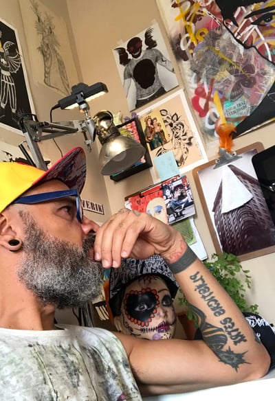 Cenário do ateliê com o artista sentado, refletindo com esboços espalhados pela mesa.