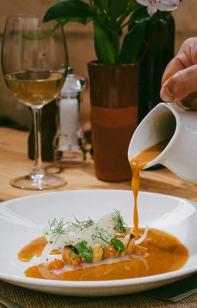 Sopa de pescado vertida en mesa con marisco, hinojo y hierbas frescas, junto a una copa de vino blanco