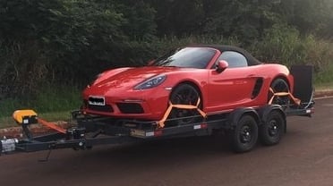 Uma carretinha tipo semi-reboque, levando um carro esportivo, vermelho, por uma estrada rural.