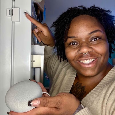 Ashley smiling while holding a smart speaker and pointing to door sensor affixed to refrigerator