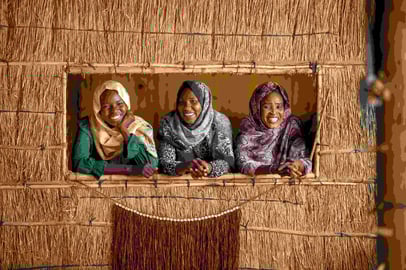 Three smiling Sudanese women looking out from a straw-framed window.