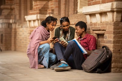 Three Sudanese students sitting and sharing content from their phones and books.