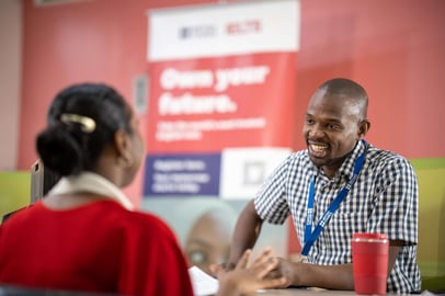 IELTS staff member smiling while talking to a test candidate at a registration desk.