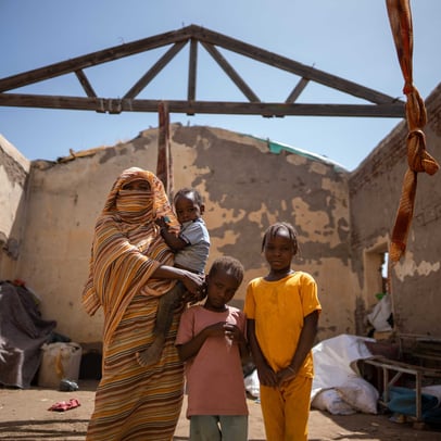 Sudanese woman and children standing inside a roofless home damaged by conflict.