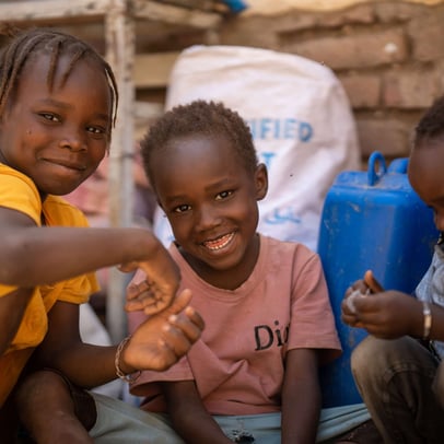 Three Sudanese children smiling and playing near water containers.