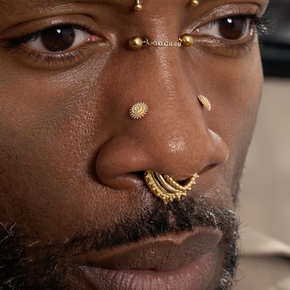 Extreme close-up of a man's face featuring an ornate gold septum ring and a chain bridge piercing