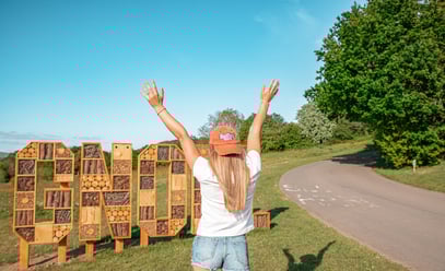 Person wearing a Big Food Festival cap standing in front of a large wooden 'GNOLL' sign with arms ra