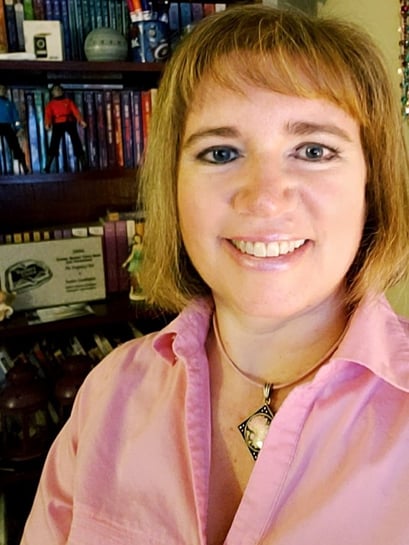 Susan Gable wearing a pink shirt, smiling, posing in front of her bookshelf.