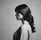 A ballet dancer is captured from behind, standing on pointe. She wears a black leotard with lace detailing on the back and a flowing white tulle skirt. Her hair is styled in a neat bun, and she is poised gracefully in a minimalist, light-filled space.