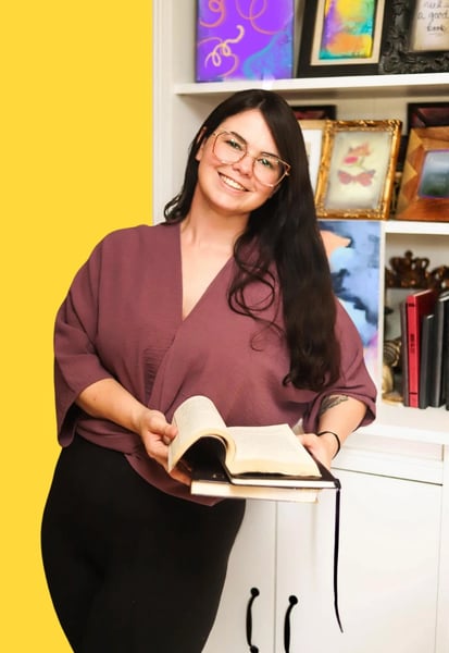 A woman stands leaning on a bookshelf with two books in her hand, one is open