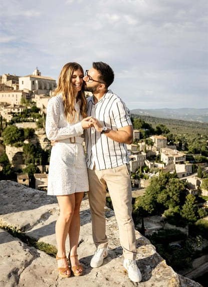 Editorial engagement photo of man kissing fiancée on cheek with scenic view of Gourdon village