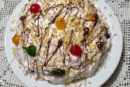 A dome shaped cake covered in whipped cream, multi-coloured glace cherries, chopped nuts and drizzled chocolate. 