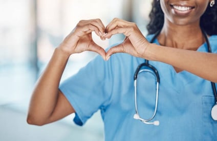 A smiling black woman in scrubs forms a heart shape with her hands, conveying warmth and compassion.