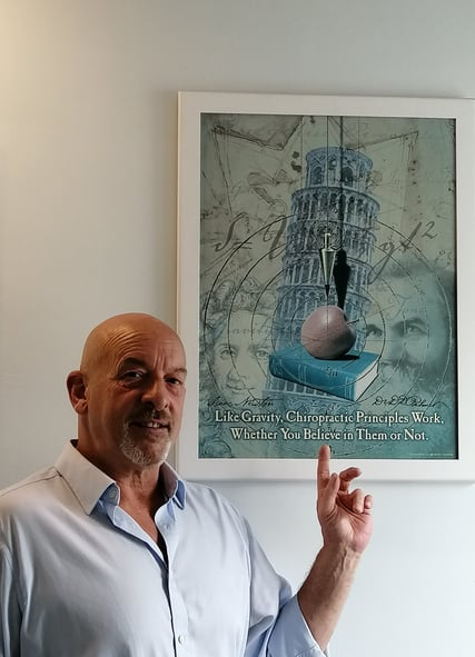 Adam Newmanin a blue shirt is standing in front of a chiropractic poster