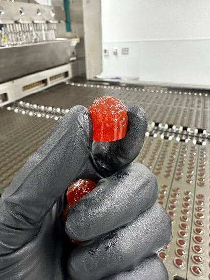 a person's hand holding a red gummy in front of automated gummy machine