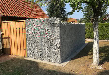 Eine als Sichtschutz zum Nachbarn errichtete Gabionenwand aus hellem Naturstein neben einem Holztor.