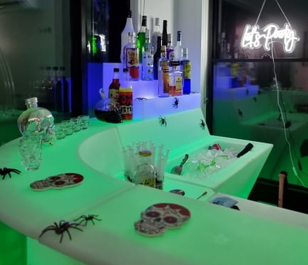 SweetAz Co. corner LED glow bar setup featuring Halloween-themed liquor, syringes, and ice, with a โLetโs Partyโ neon sign gl