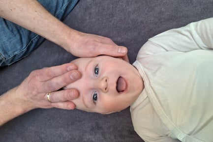 een baby ligt op zijn rug en lacht terwijl 2handen zijn hoofd vasthouden.