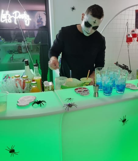 Bartender in Halloween makeup preparing drinks at a glowing green LED bar decorated with crawling spiders.