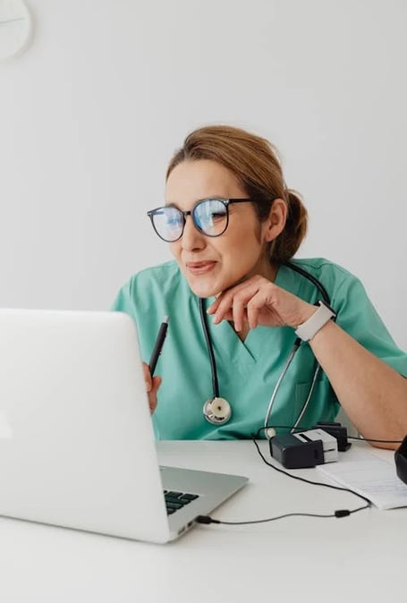 a woman in a green scrubsuit and glasses is looking at a laptop