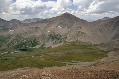 Expansive view of a rugged mountain range with alpine meadows and a small lake below.