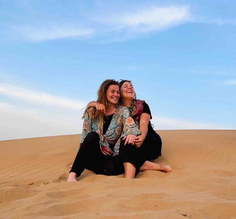 Eva and Manon, two travelers sitting in the Jaisalmer desert, surrounded by golden dunes at sunset.