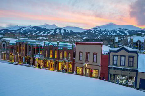 Snow-covered street lined with colorful historic buildings at sunset with ski slopes behind.