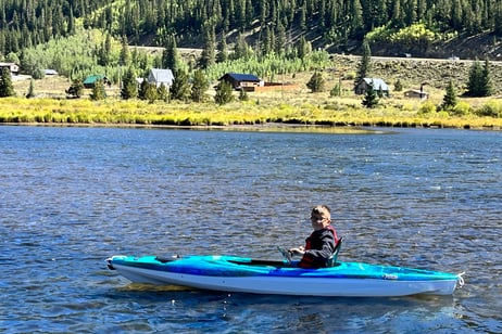 Young boy paddling a bright blue kayak on a calm river with forest and cabins in the background.
