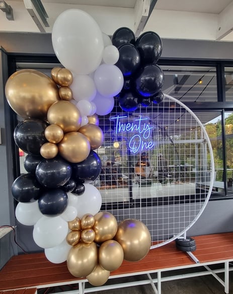 White circle mesh backdrop with blue Twenty One neon sign and a gold, black and white balloon garland, Redlands